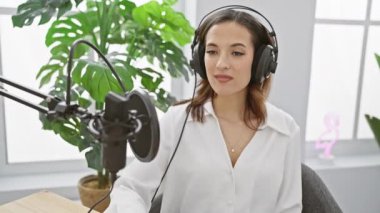 Genç bir İspanyol kadın, modern bir stüdyoda kulaklık ve mikrofonla podcast yapıyor..