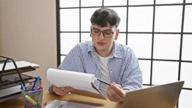 Çizgili gömlekli odaklanmış genç bir adam iyi organize edilmiş bir ofis masasında evrak okuyor.