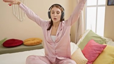 Pembe pijama setiyle, rahat bir yatak odasında kulaklıkla müzik dinleyen genç bir kadın..