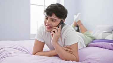 Neşeli genç bir adam, aydınlık bir yatak odasında, bir yatakta dinlenirken, akıllı bir telefonla konuşuyor, rahat ve modern bir yaşam tarzı yayıyor..