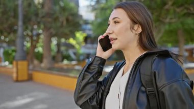 Genç esmer bir kadın, güneşli bir şehir sokağında yürürken telefonuyla konuşuyor, günlük şehir hayatını somutlaştırıyor..