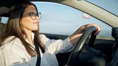 Genç, güzel, İspanyol bir kadın. Araba kullanıyor. Gözlüklü, gülümsüyor.