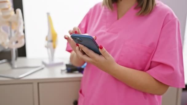 Leende Kvinna Rosa Skrubb Bläddrar Sin Telefon Klinik Med Ryggrad — Stockvideo