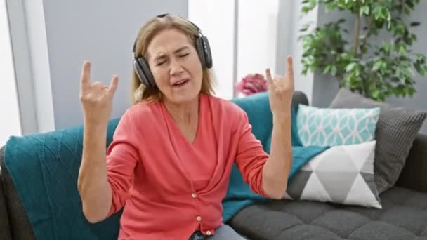 金发老妇人喜欢家里带着耳机的音乐 在舒适的客厅里展示摇滚手势 — 图库视频影像