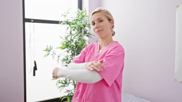 Confident Blonde Woman Pink Scrubs Arms Crossed Bright Clinic Interior — Stock Video