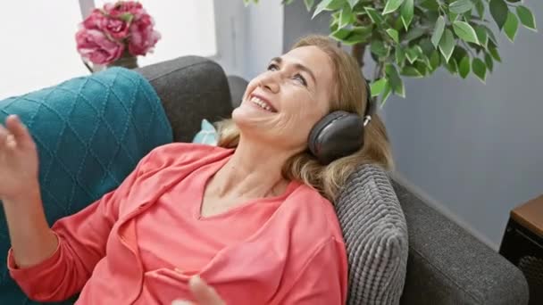 一位快乐的中年妇女在耳机里听着音乐 舒舒服服地躺在沙发上 她的笑声暗示着幸福 — 图库视频影像