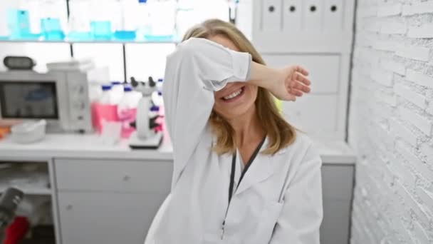 陽気な中年ブロンド女性科学者は楽しんで ラボでピークを演奏し 輝く笑顔で楽しい顔を示しています — ストック動画