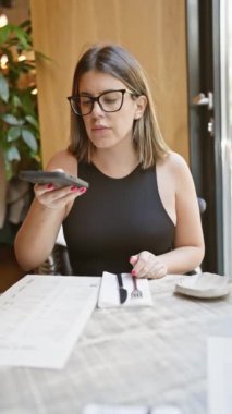 Gözlüklü ve siyah elbiseli genç bir İspanyol kadın kafeterya masasında menüsü olan akıllı telefonunu kullanıyor..