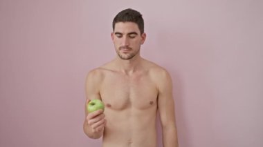 Tişörtsüz İspanyol genç adam, gülümseyen, mutluluğun tadını çıkaran, çapraz kollu bir elma tutan, serin pembe bir arka plan üzerinde sağlık ve pozitiflik bir resim.