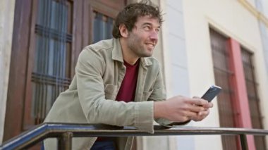 Akıllı telefon tutan neşeli bir adam güneşli şehir ortamında korkuluklara yaslanır.