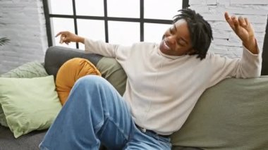 Genç bir Afro-Amerikalı kadın. Rasta bukleleriyle evdeki rahat oturma odasında neşeyle dans ediyor. Etrafı renkli yastıklar ve kanepelerle çevrili..