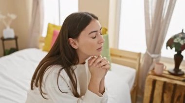 Düşünceli, genç bir İspanyol kadın aydınlık, rahat yatak odasında dua ediyor, huzur ve maneviyat hissi yayıyor..