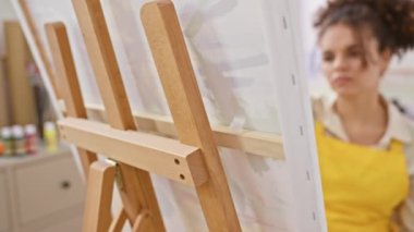 Ciddi yüz ifadesi olan, kendine güvenen kıvırcık saçlı kadın sanatçı stüdyoda doğal olarak oturuyor.