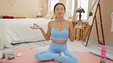 Rahat genç Çinli kadın yatak odasında huzur buluyor, yoga minderinde gözleri kapalı gülümsüyor, meditasyon jestleri yapıyor.