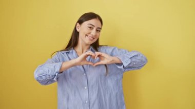 Neşeli genç İspanyol kadın, sarı izole bir arka planda duruyor, gömlek giyiyor, ellerini kullanarak bir aşk-kalbi sembolü yaratıyor. Güzellik ve neşe dolu romantik bir konsept..