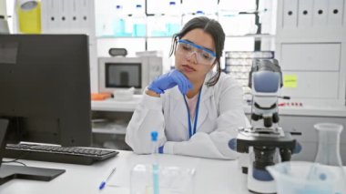 Mikroskop, bilgisayar ve laboratuvar ekipmanları olan düşünceli bir kadın bilim adamı..