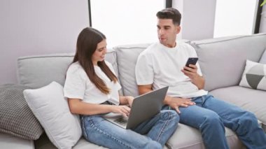 İyi ışıklandırılmış bir oturma odasında gri bir koltukta oturan bir kadın ve bir erkek, akıllı bir telefon tutuyor ve kadın dizüstü bilgisayar kullanıyor..