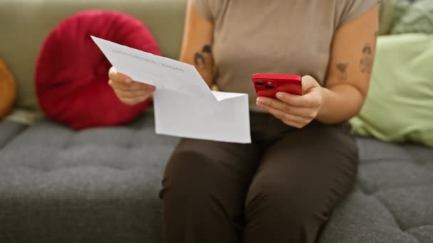 一位年轻的惊慌失措的女人坐在屋里看书 同时在客厅里拿着智能手机 — 图库视频影像