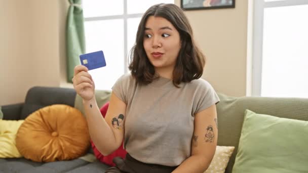 年轻而美丽的惊慌失措的女人在家里尽情地享受着信用卡的成功 愉快地坐在沙发上 用手指指点着出色的象征 流露出自信和积极向上 — 图库视频影像