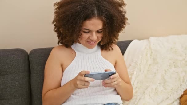 一位卷发的年轻女子在屋里沙发上享受着智能手机 流露出喜悦和放松 — 图库视频影像