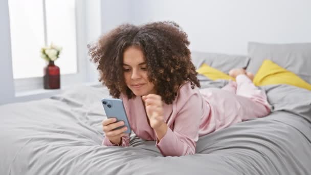 ベッドの上に横たわる 楽しい カーリーな髪のヒスパニックの女性は 自信を持って笑顔で片側を指しています 寝室のシーンでスマートフォンでテキストメッセージングに従事 — ストック動画