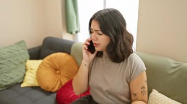 Endişeli genç bir İspanyol kadın renkli bir oturma odasında yastıklarla konuşuyor..