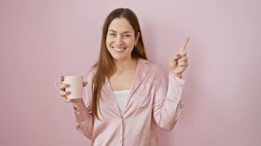 Mavi gözlü neşeli genç bir kadın, sabah kahve fincanını sallarken mutlu bir şekilde yan tarafı gösteriyor, izole edilmiş, dostane pembe arka planda rahat pijamalarıyla..