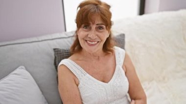 Neşeli orta yaşlı kadın, kendine güvenen kızıl saçlı, olumlu duygularını ifade eden, başparmağını kaldırmış, kanepeye oturmuş elbise giymiş, evdeki rahat oturma odasında ağzı açık bir şekilde ışıldıyor.