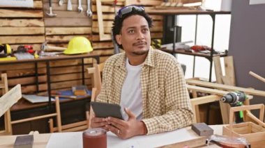 Kıvırcık saçlı genç adam ahşap atölyesinde tablet kullanıyor. Etrafı aletler ve güvenlik ekipmanlarıyla çevrili.