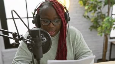 Saçları örgülü Afrikalı Amerikalı bir kadın radyo stüdyosunda senaryo okuyor..