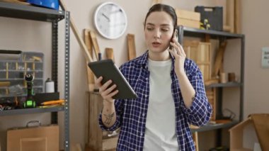 Bir marangozluk atölyesinin organize kaosunun ortasında bir tablet ve bir telefon görüşmesi ile odaklanmış genç bir kadın,.