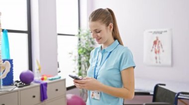 Üniformalı, gülümseyen kadın terapist. Parlak bir rehabilitasyon merkezinde tıbbi ekipmanla akıllı telefon kullanıyor.