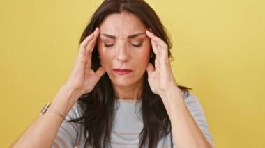 Gergin, olgun İspanyol kadın izole edilmiş sarı arka planda duruyor, elleri başının üstünde, migren baş ağrısını ifade eden üzgün bir yüz. Ağrısı çaresizce acı çektiğini gösteriyor..