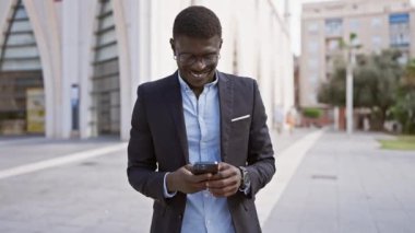 Güneşli bir caddede akıllı telefon kullanan şık bir adam, şehir yaşam tarzını ve bağlantıyı çağrıştırıyor..