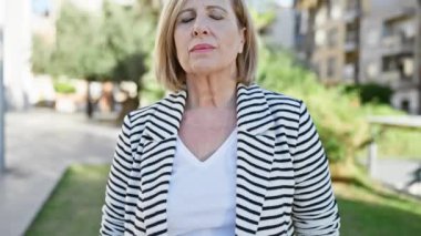 Olgun sarışın kadın, gözleri kapalı güneşli bir parkta huzur içinde duruyor.