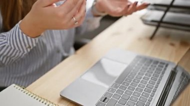 Genç bir kadın, ofis ortamında bir iş görüşmesi sırasında dizüstü bilgisayar ve not defteriyle el hareketleriyle iletişim kuruyor..