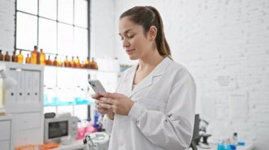 Beyaz laboratuvar önlüklü profesyonel bir kadın, parlak, modern bir laboratuvar ortamında akıllı telefon kullanıyor. Sağlık hizmetlerinde teknolojiyi ön plana çıkarıyor..
