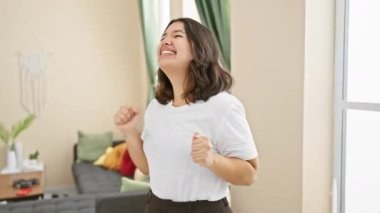 Heyecanlı genç İspanyol kadın zaferi evde kutluyor! Kollar havada, güzel yüzle oturma odasında zaferi sevinçle ifade eden bir yaşam tarzı zaferi! Evet.!