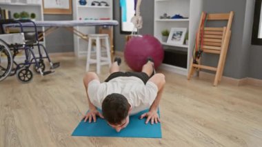 İspanyol adam yoga minderi, tekerlekli sandalye ve fizyoterapi ekipmanıyla rehabilitasyon kliniğinde egzersiz yapıyor..