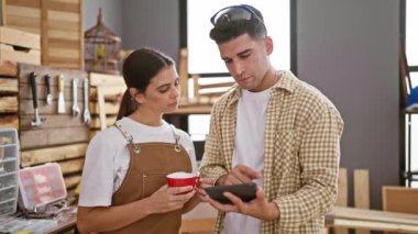 Atölyedeki erkek ve kadın, arka planda ahşap işleme araçlarıyla tablet üzerindeki tasarımları gözden geçiriyorlar..