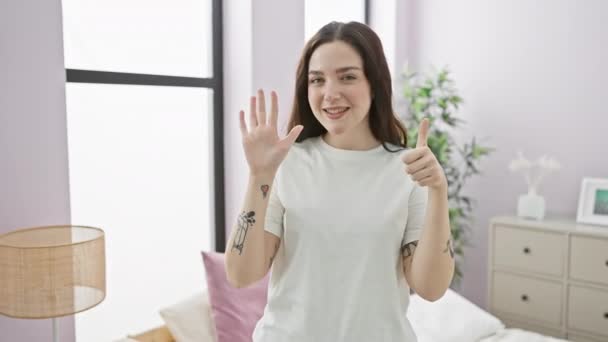 一个快乐的年轻女人 穿着睡衣坐在卧室的床上 一边举起手 一边笑着 一边用手指指点着第六个积极的概念 — 图库视频影像