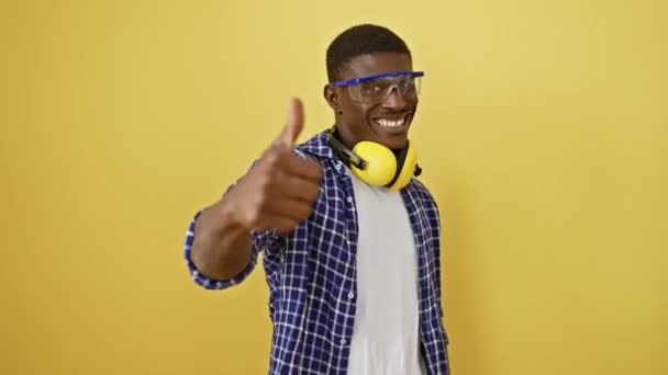 アフリカ系アメリカ人男性は 安全眼鏡で 自信を持って立って 彼の笑顔で明らかに優れた承認サインを点滅させます 活気に満ちた黄色の背景から隔離された彼は成功を体現します — ストック動画