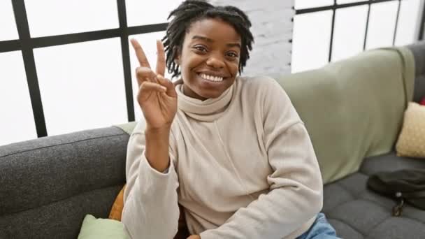 快乐的年轻黑人女人 头发蓬乱 坐在舒适的沙发上 两手空空地展示胜利的标志 用积极的目光看着相机 在家里的室内画像 — 图库视频影像