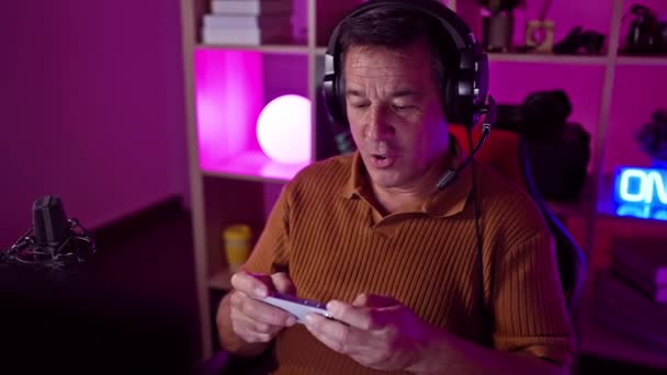 一个带着耳机的成熟男人晚上在霓虹灯下的游戏室里使用智能手机 传达一种现代高科技的生活方式 — 图库视频影像