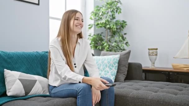 一个穿着休闲装的快乐的高加索女人在一间现代客厅里的一个闷热的沙发上放松下来 手里拿着一个遥控器 — 图库视频影像