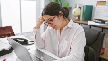 Odaklanmış İspanyol bir kadın, parlak bir ofis ortamında dizüstü bilgisayarının başında çalışarak profesyonellik ve adanmışlığı aktarıyor..