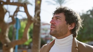 Sakallı mutlu bir İspanyol adam güneşli bir parkta sıcacık gülümsüyor, günlük şehir hayatını canlandırıyor..