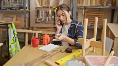 Odaklanmış İspanyol bir kadın, kapalı alandaki marangozluk atölyesinde detaylara dikkat çekerken telefonla konuşuyor..