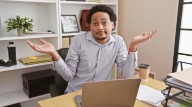 Kafası karışmış, afro-amerikan bir iş adamı, ofis masasında hiçbir şey bilmeden oturuyor, elleri ve kolları havada şaşkın bir yüz ifadesi sergiliyor. İş yerindeki şüpheyi somutlaştıran.