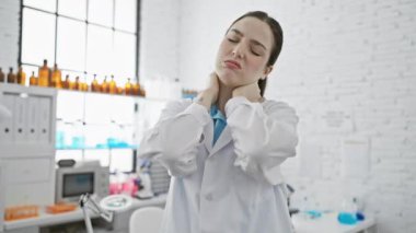 Laboratuvar önlüğü giymiş stresli bir kadın bilim adamı klinik laboratuvar ortamında boyun ağrısı çekiyor..